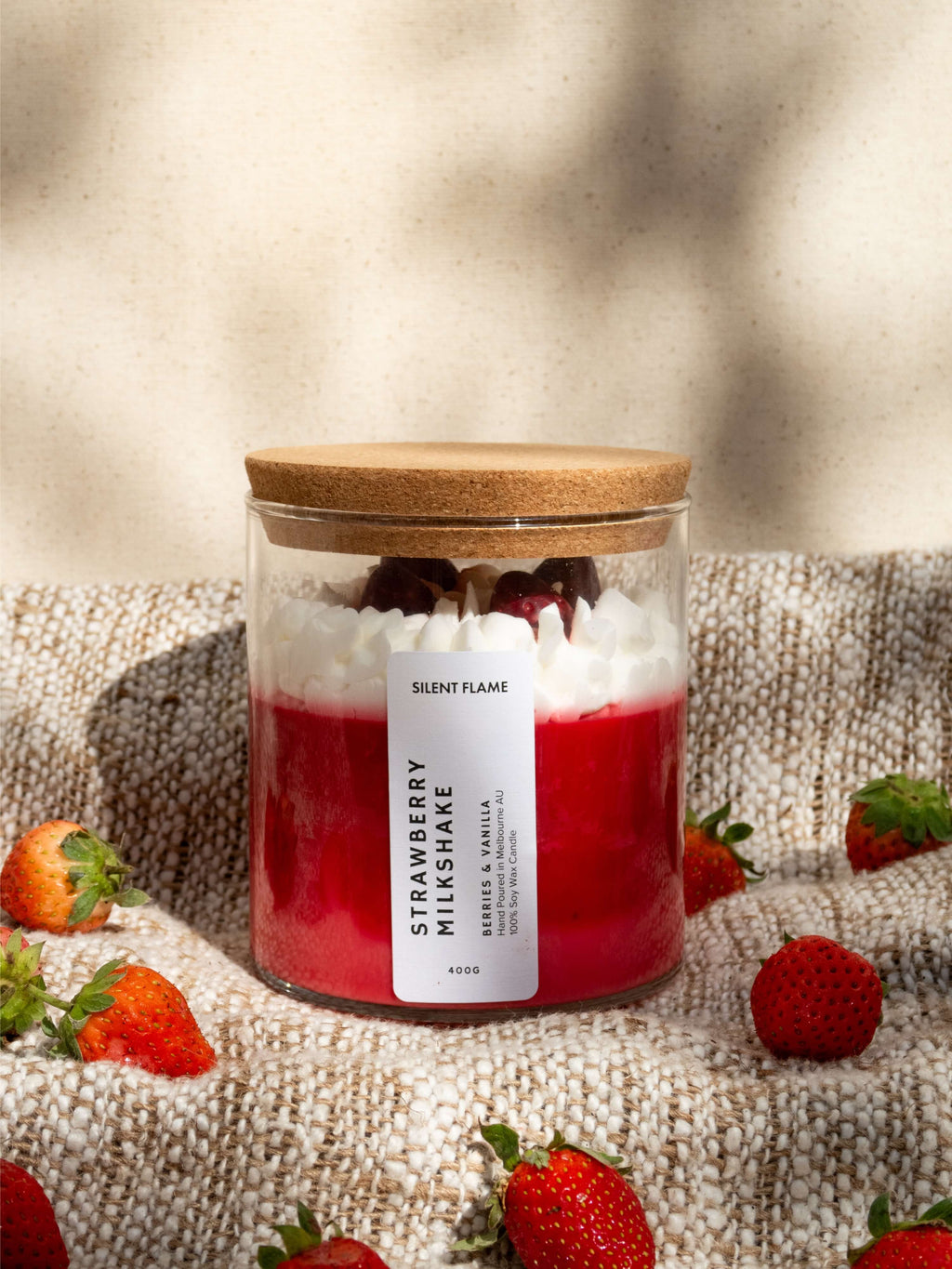 Glass jar with wooden lid containing a strawberry milkshake candle, surrounded by strawberries on a textured surface.